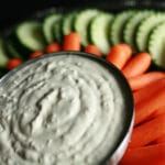 A bowl of wasabi ginger dip, surrounded by cucumber slices and baby carrots.
