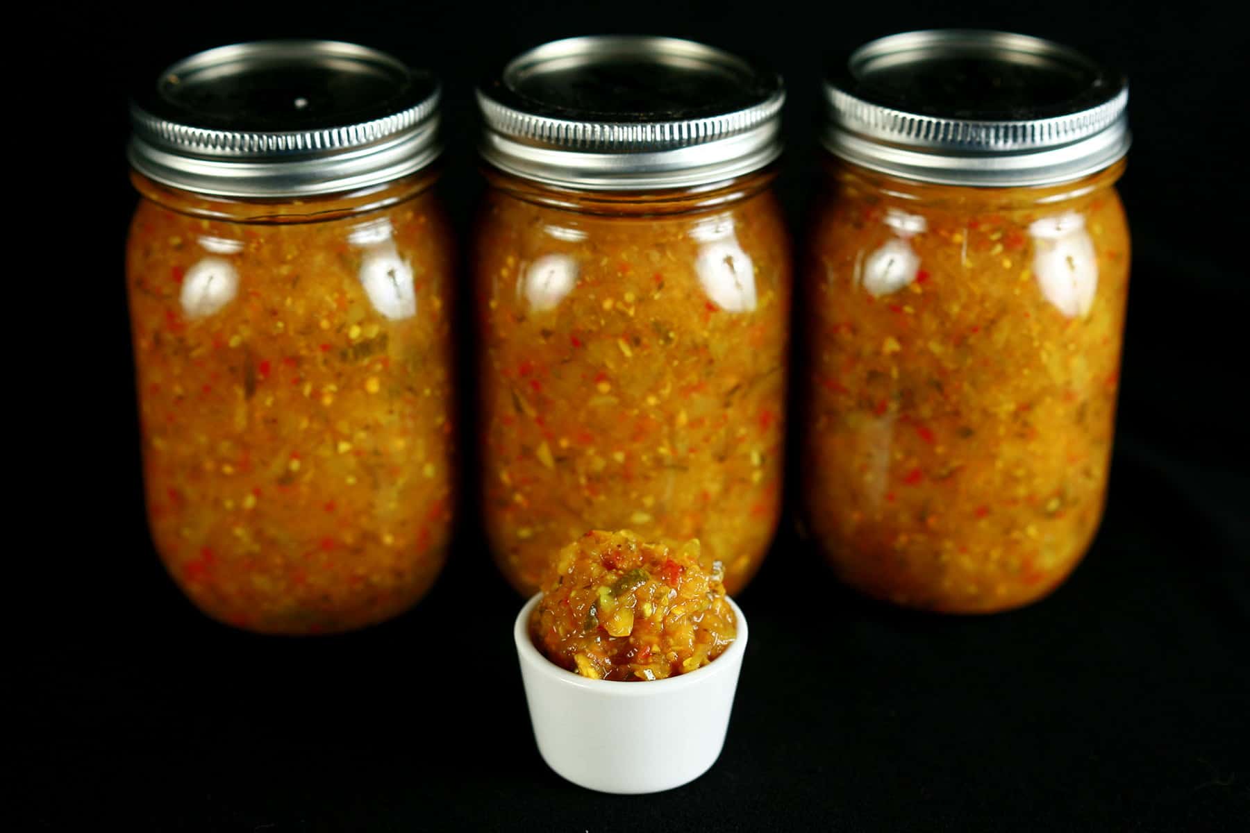 3 jars of Hoppy Dill Pickle Relish. It's a golden orange relish with flecks of dark green and red throughout. There is a small cup of the relish in front of the jars.