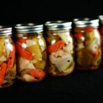 A line of 5 jars of mixed vegetable pickles on a black background. Baby carrots, broccoli, onion, celery, cauliflower, and jalapeno are all visible.