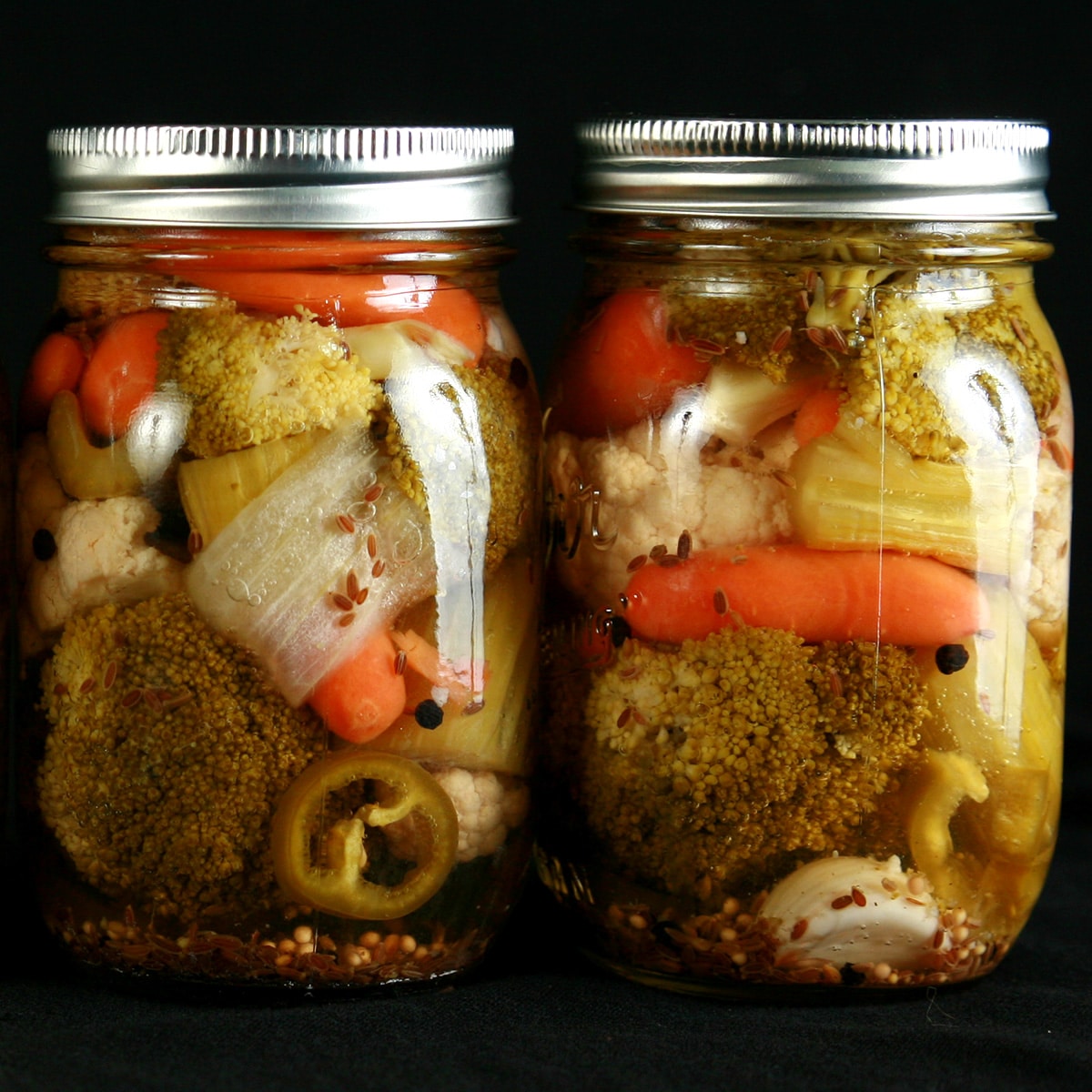 2 jars of mixed vegetable pickles on a black background. Baby carrots, broccoli, onion, celery, cauliflower, and jalapeno are all visible.