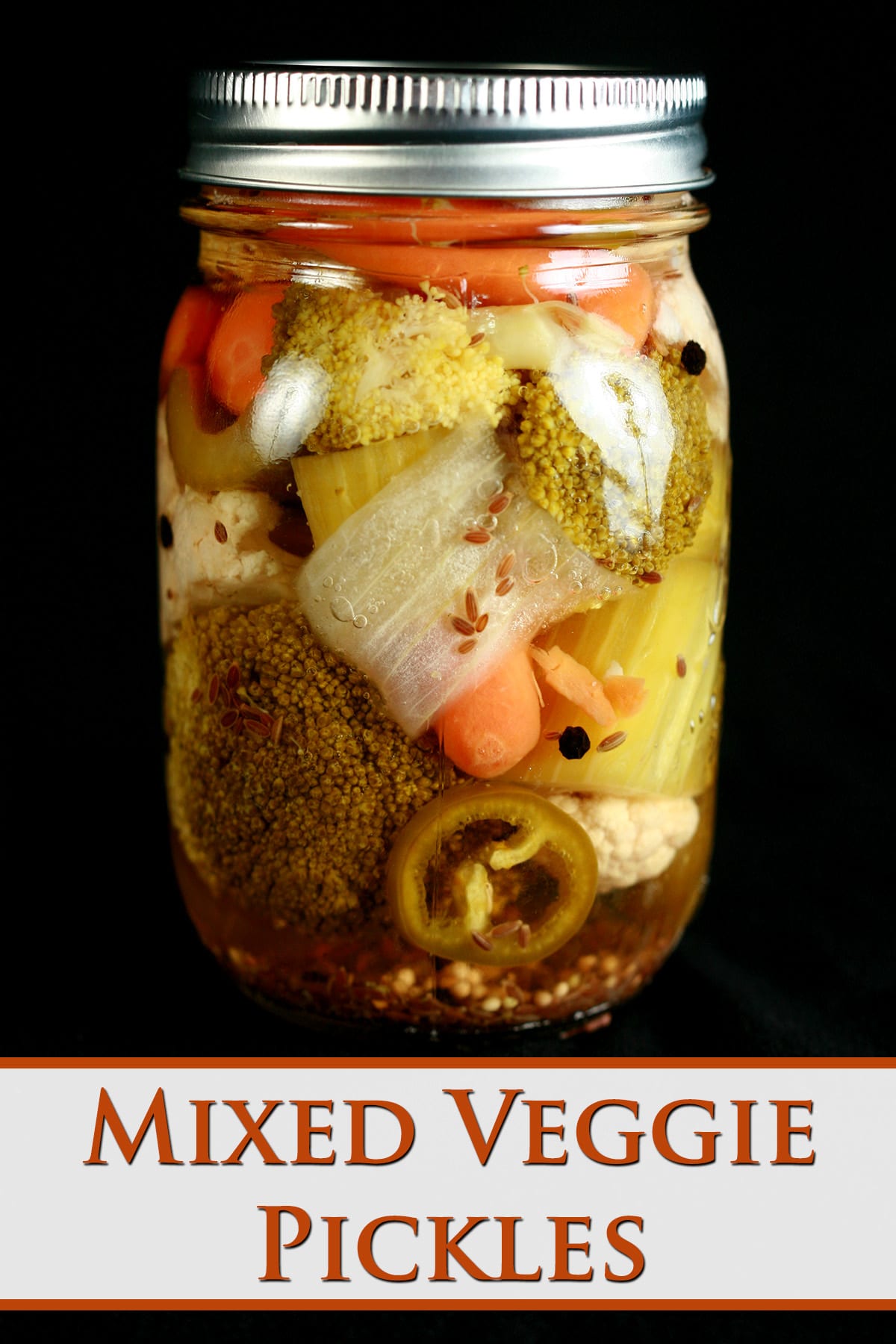 A jar of mixed vegetable pickles on a black background. Baby carrots, broccoli, onion, celery, cauliflower, and jalapeno are all visible.