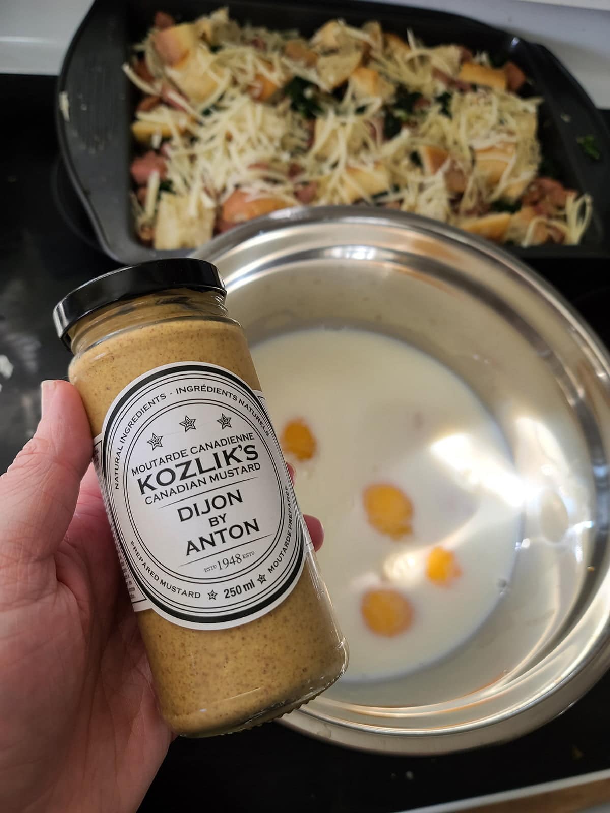 A hand holding a jar of Kozlik's Dijon mustard above a mixing bowl with milk and eggs in it.