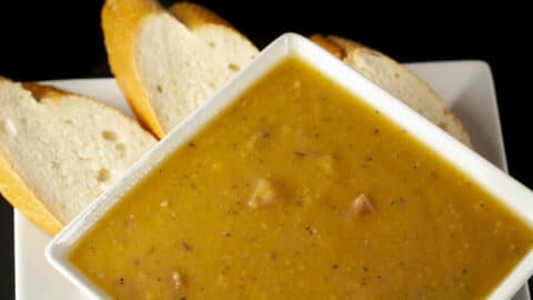 A white, square shaped bowl of smooth French Canadian Pea Soup, with sliced baguette next to it.