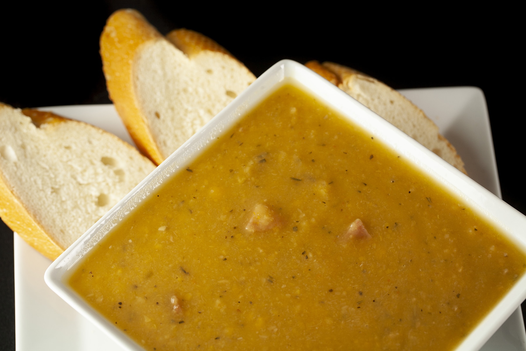 A white, square shaped bowl of smooth French Canadian Pea Soup, with sliced baguette next to it.