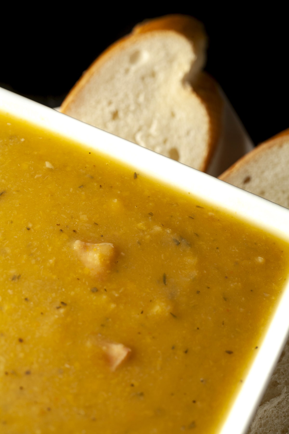 A white, square shaped bowl of smooth French Canadian Pea Soup, with sliced baguette next to it.