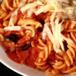 A bowl of pasta in red sauce, with bits of pepperoni and sliced black olives visible.