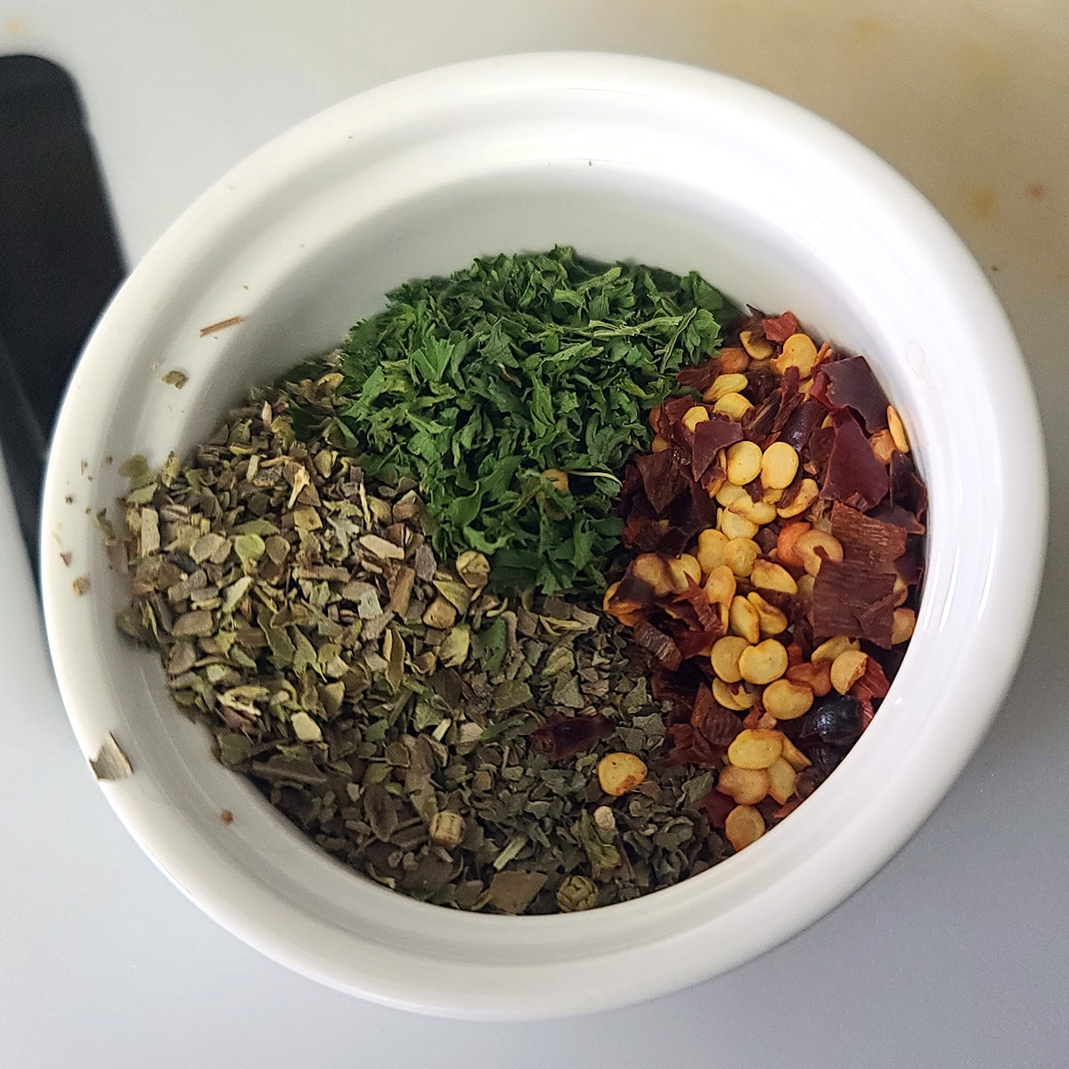 A small ramekin with dried green herbs and pepper flakes in it.