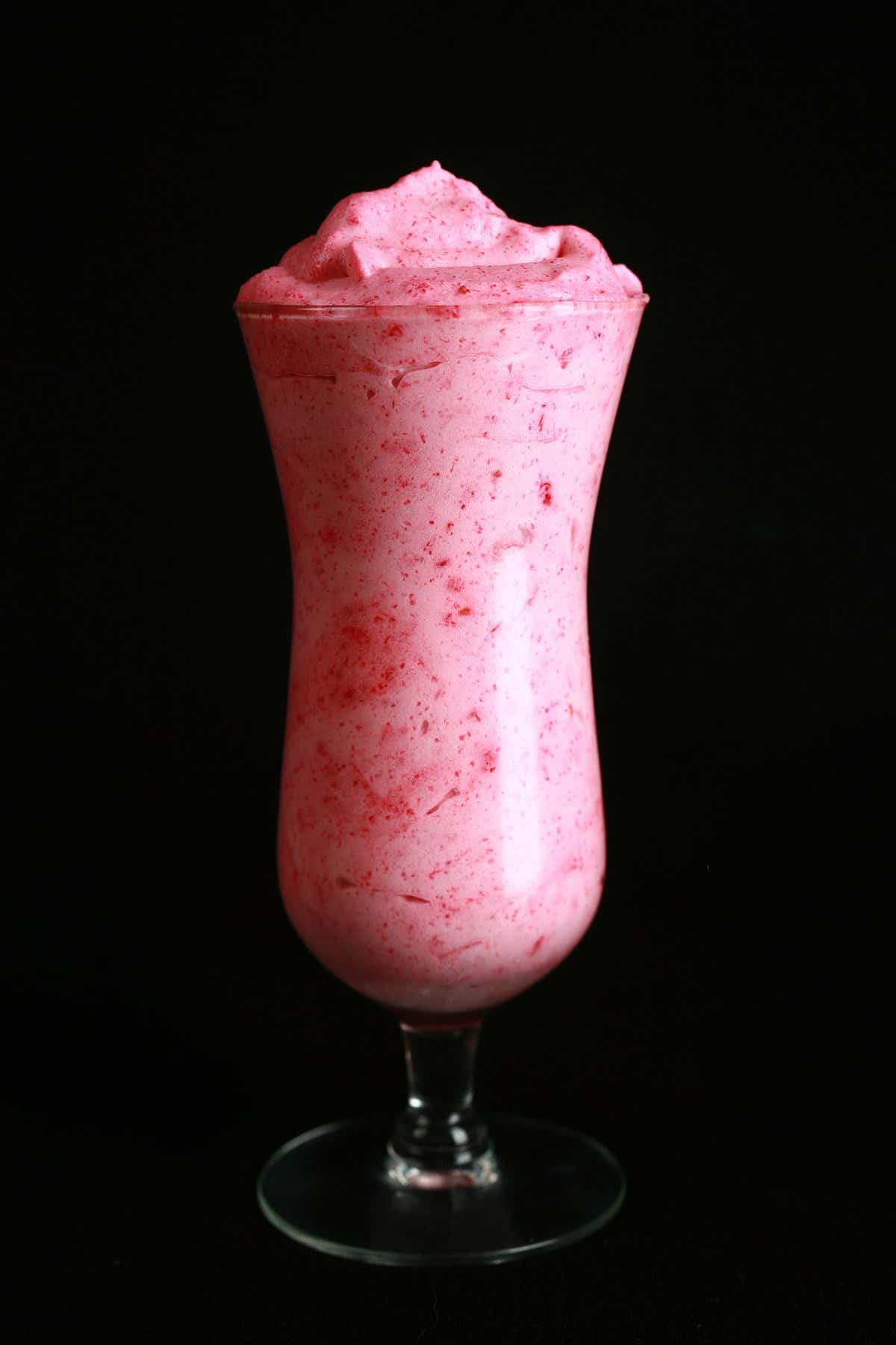 A tall, tulip shaped cocktail glass is filled with a deep pink raspberry mousse, against a black background,