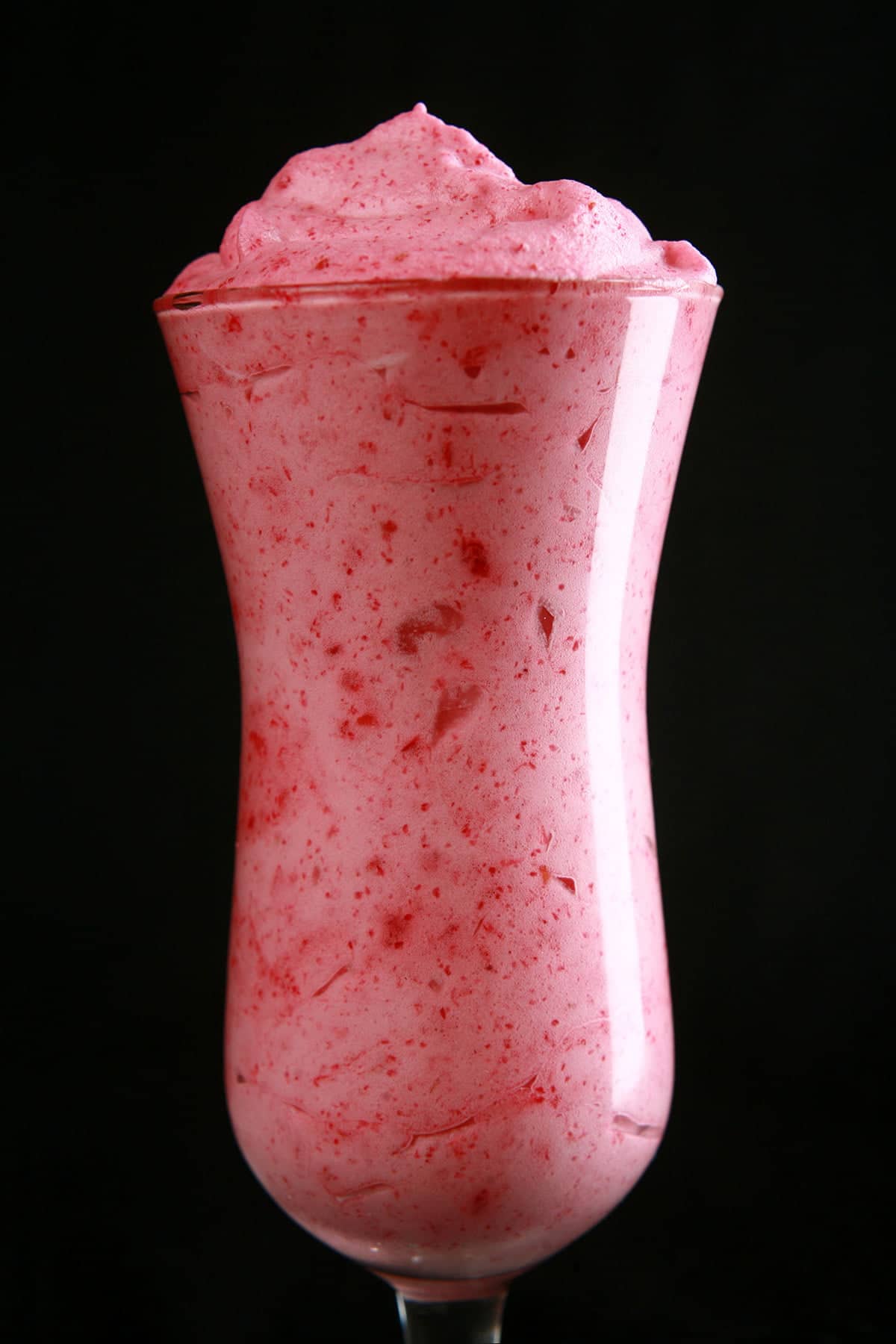 A tall, tulip shaped cocktail glass isfilled with a deep pink raspberry mousse, against a black background,