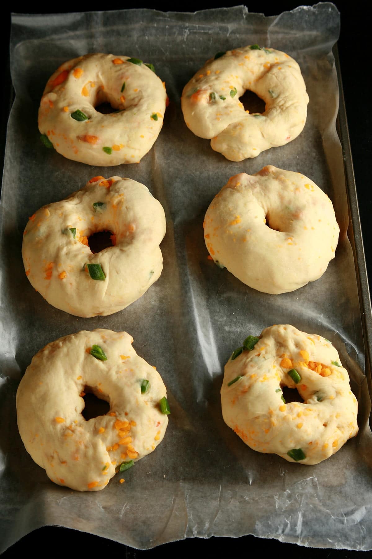6 raw bagels are shown on a parchment lined surface. They are generously studded with jalapeno pepper pieces and cheese.