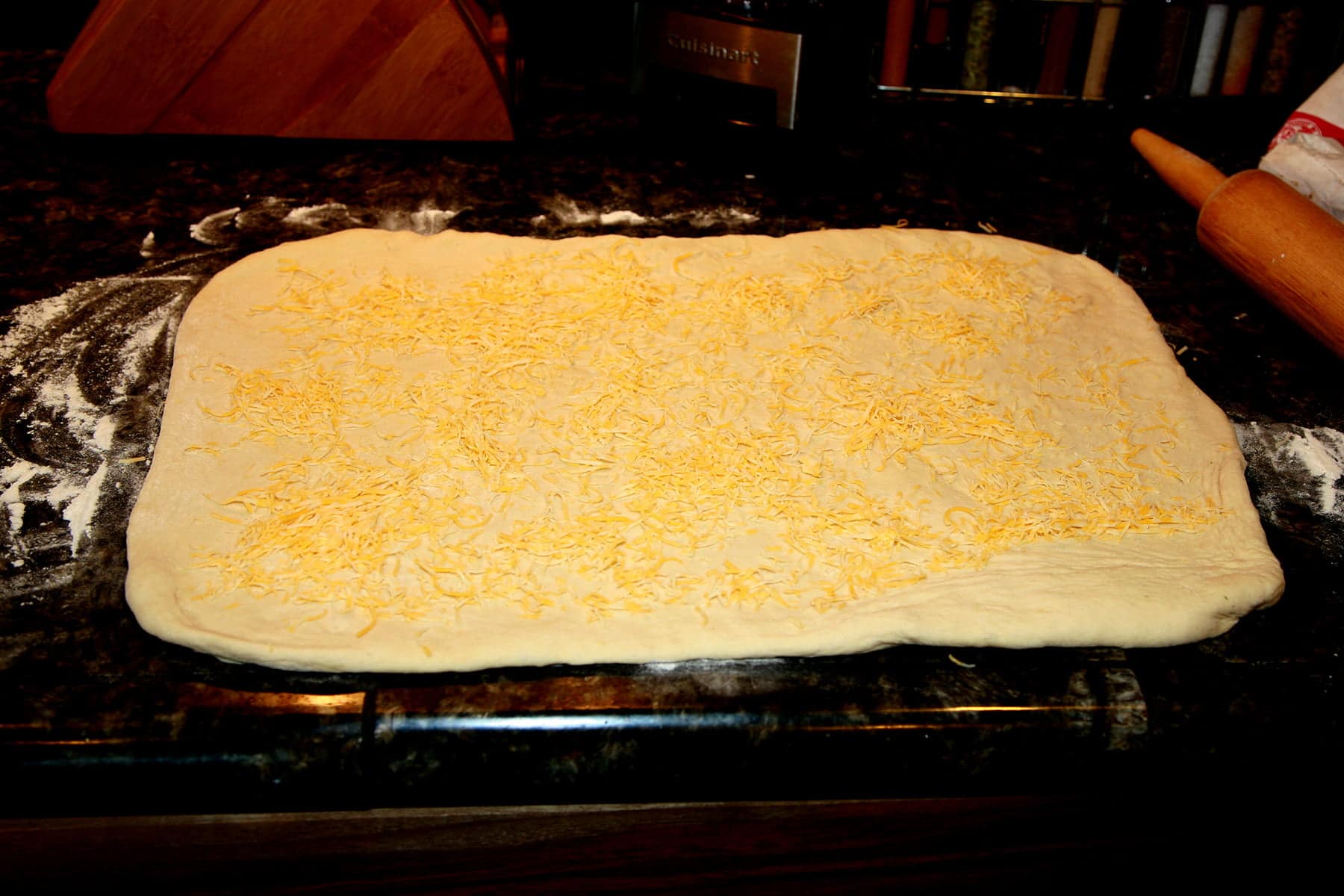 A rectangle of dough is rolled out on a counter, and covered in shredded cheese.