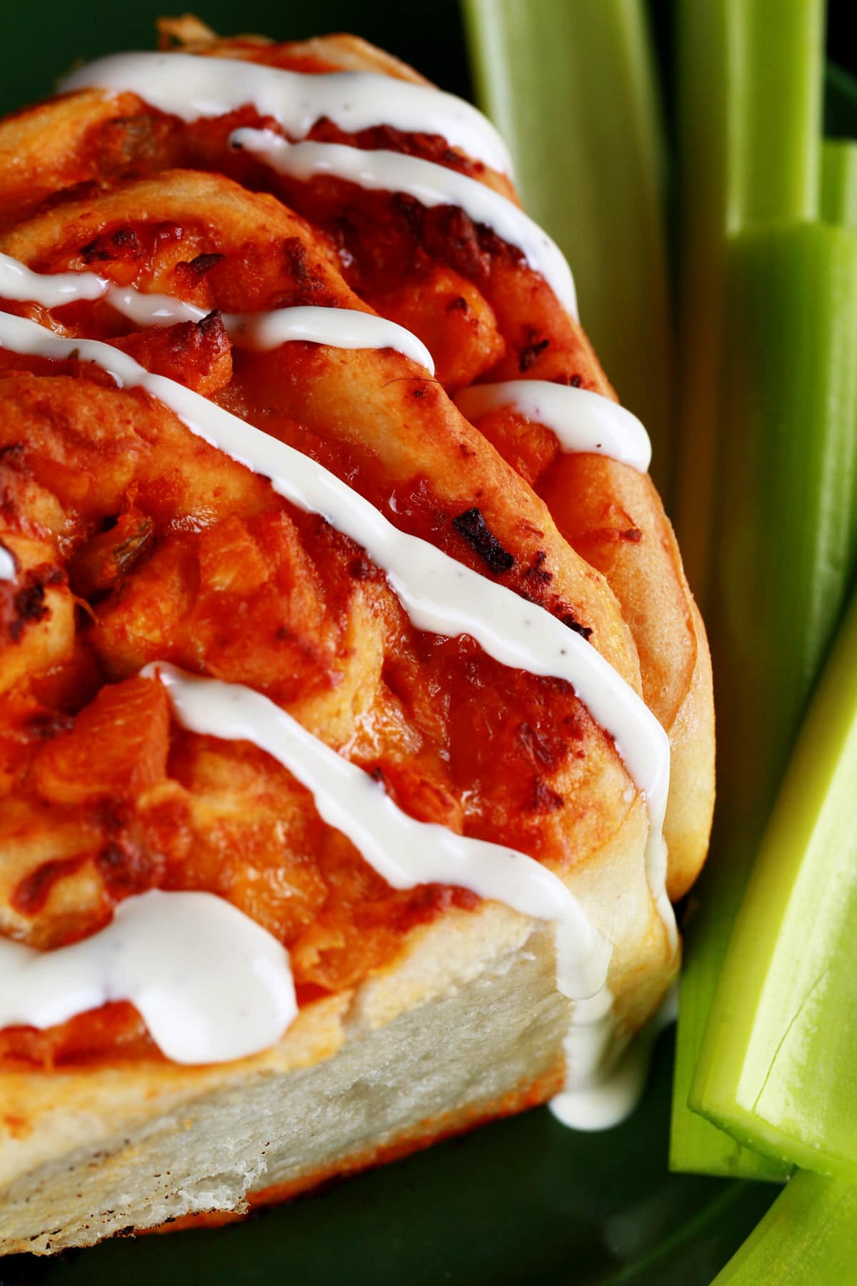 A close up photo of a buffalo chicken bun - like a cinnamon bun, but with a buffalo chicken filling instead. It is drizzled with ranch dressing, and has celery sticks on the side.