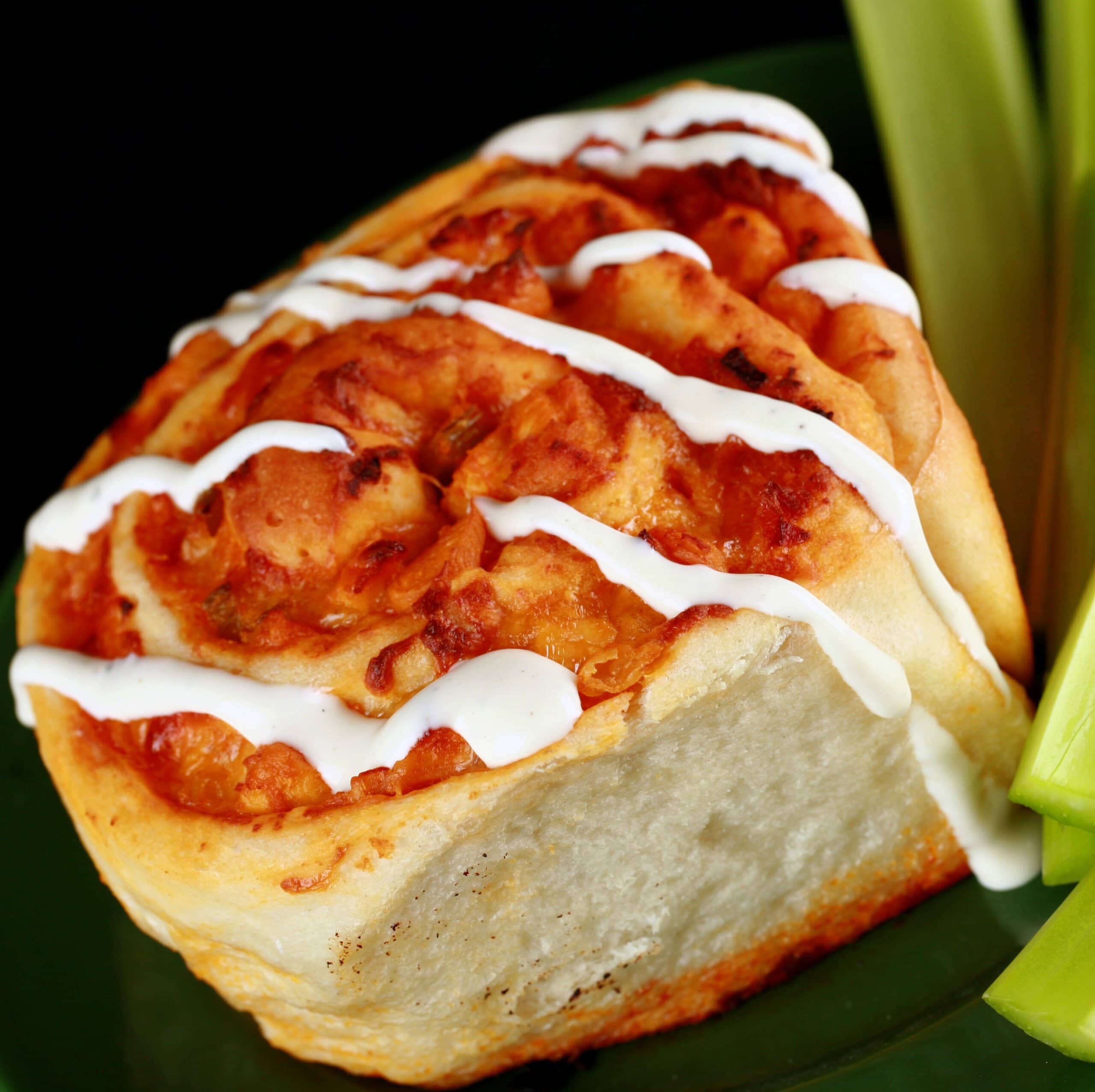 A close up photo of a buffalo chicken roll - like a cinnamon bun, but with a buffalo chicken filling instead. It is drizzled with ranch dressing, and has celery sticks on the side.