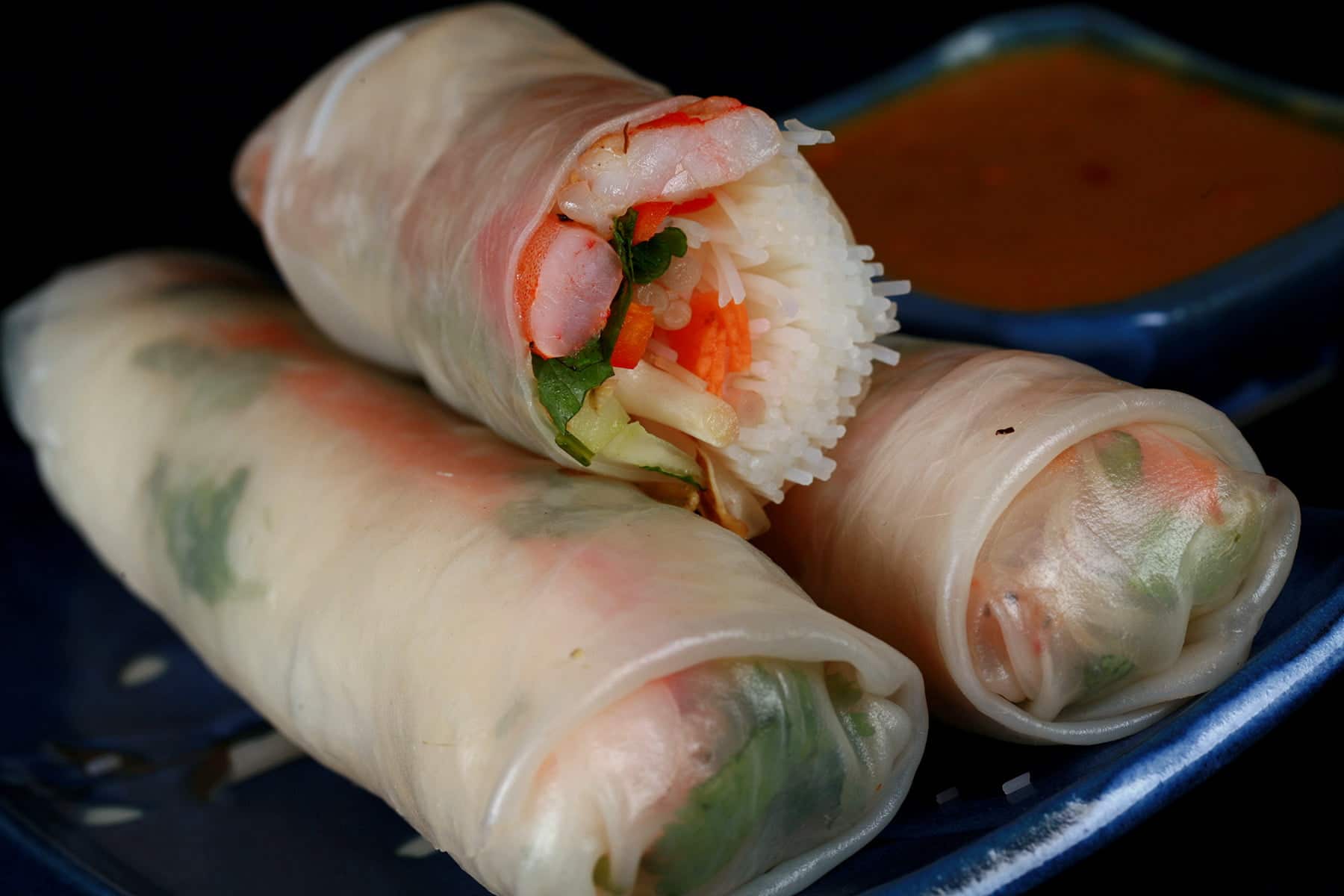 2 and a half curried shrimp spring rolls are arranged on a blue plate, with a small bowl of spicy peanut dip next to it.