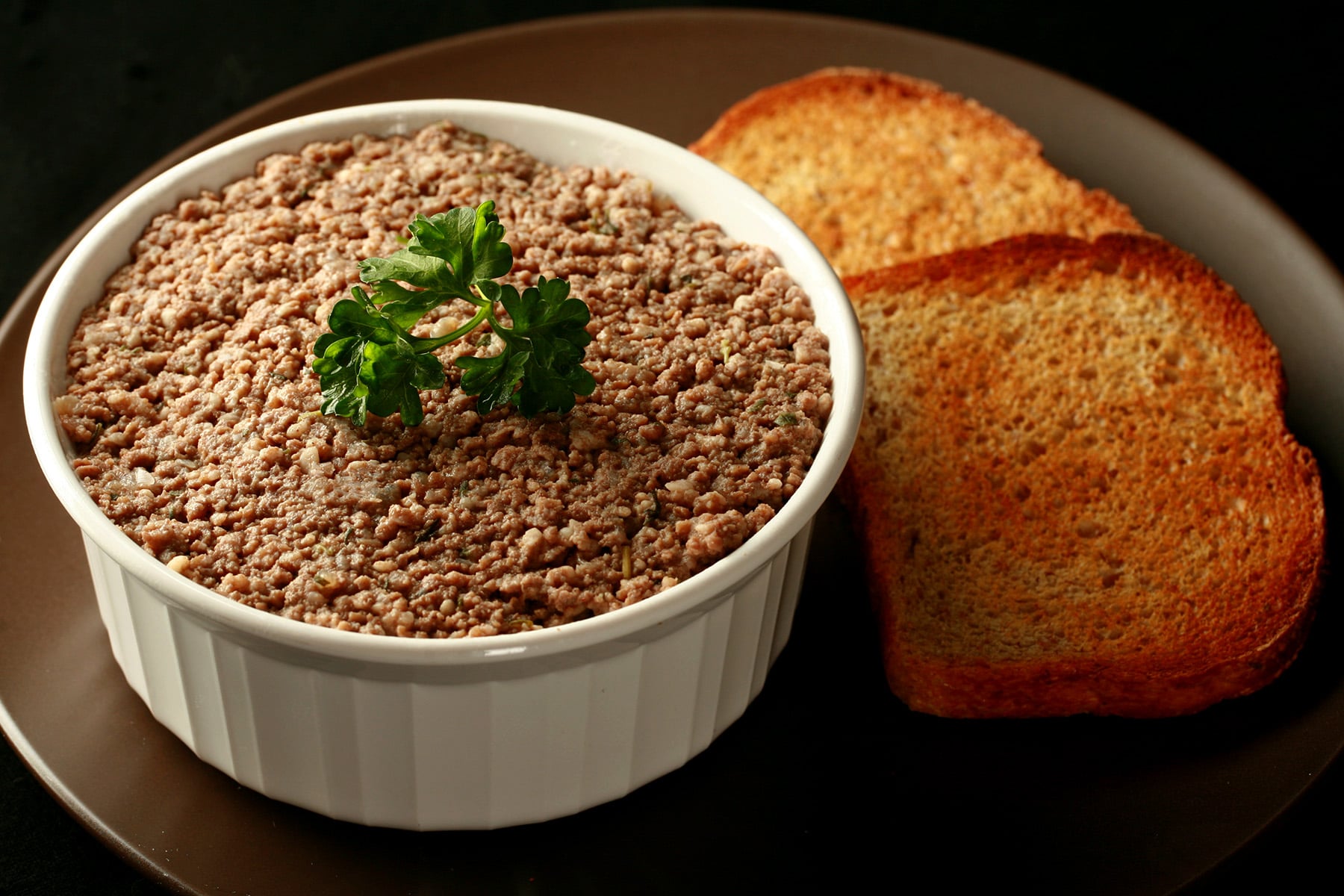 A pot of cretons - a French Canadian pork pate - in a small white ramekin, next to slices of toast.