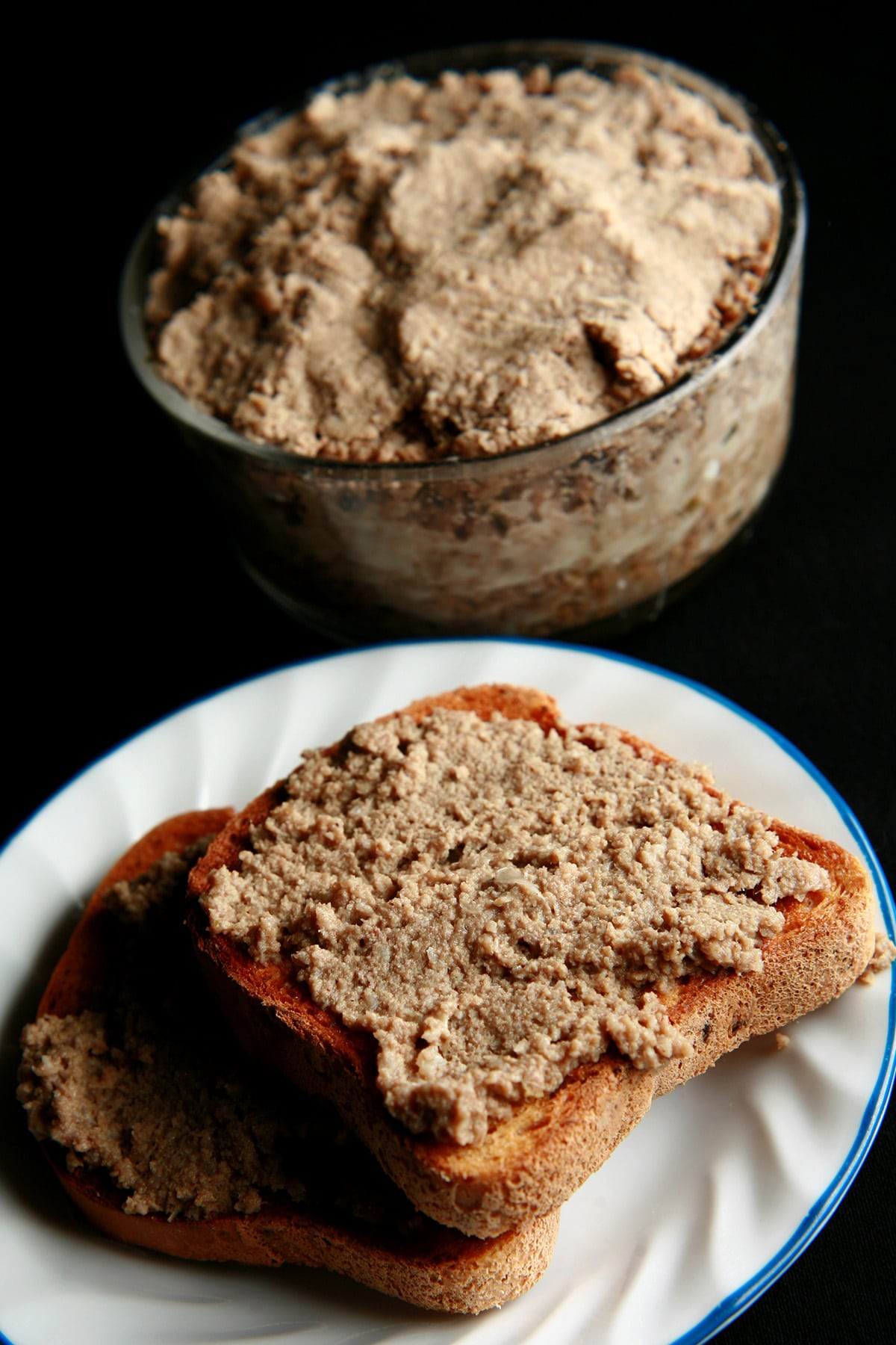 A slice of toast spread with cretons, on a small white plate.  There is a glass dish packed with cretons next to it.