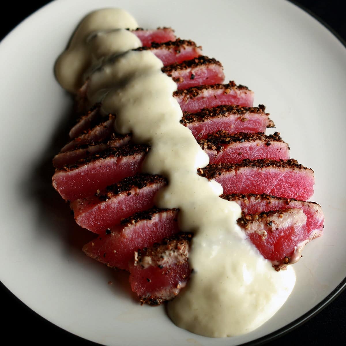 A pepper crusted tuna steak - rare - that has been sliced up, fanned out on a small white plate, and drizzled with wasabi cream sauce.