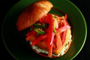 A very close up view of a paneer burger - a pakora battered patty of paneer, with cilantro-mint mayonnaise and a pickled slaw made of red peppers, carrots, and jalapenos, on a bun.