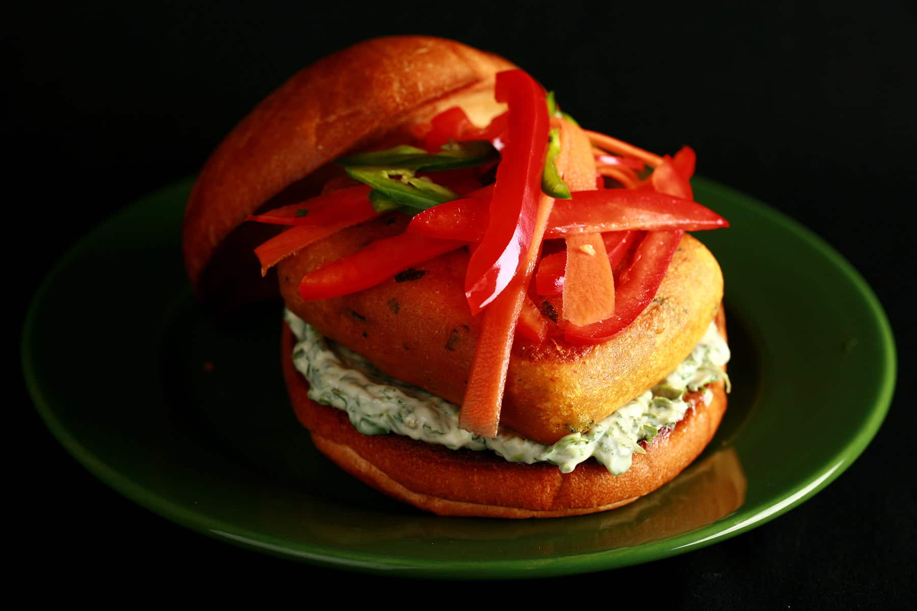 A very close up view of a paneer burger - a pakora battered patty of paneer, with cilantro-mint mayonnaise and a pickled slaw made of red peppers, carrots, and jalapenos, on a bun.