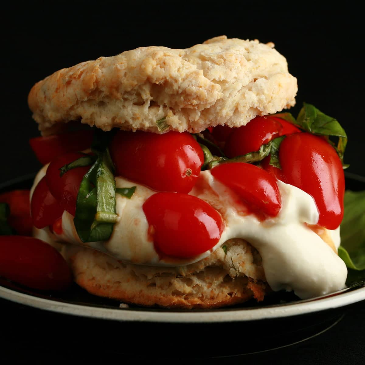 A baking powder biscuit "sandwich" on a small black plate.  The biscuit has soft white cheese, cherry tomatoes, and fresh basil as its filling.