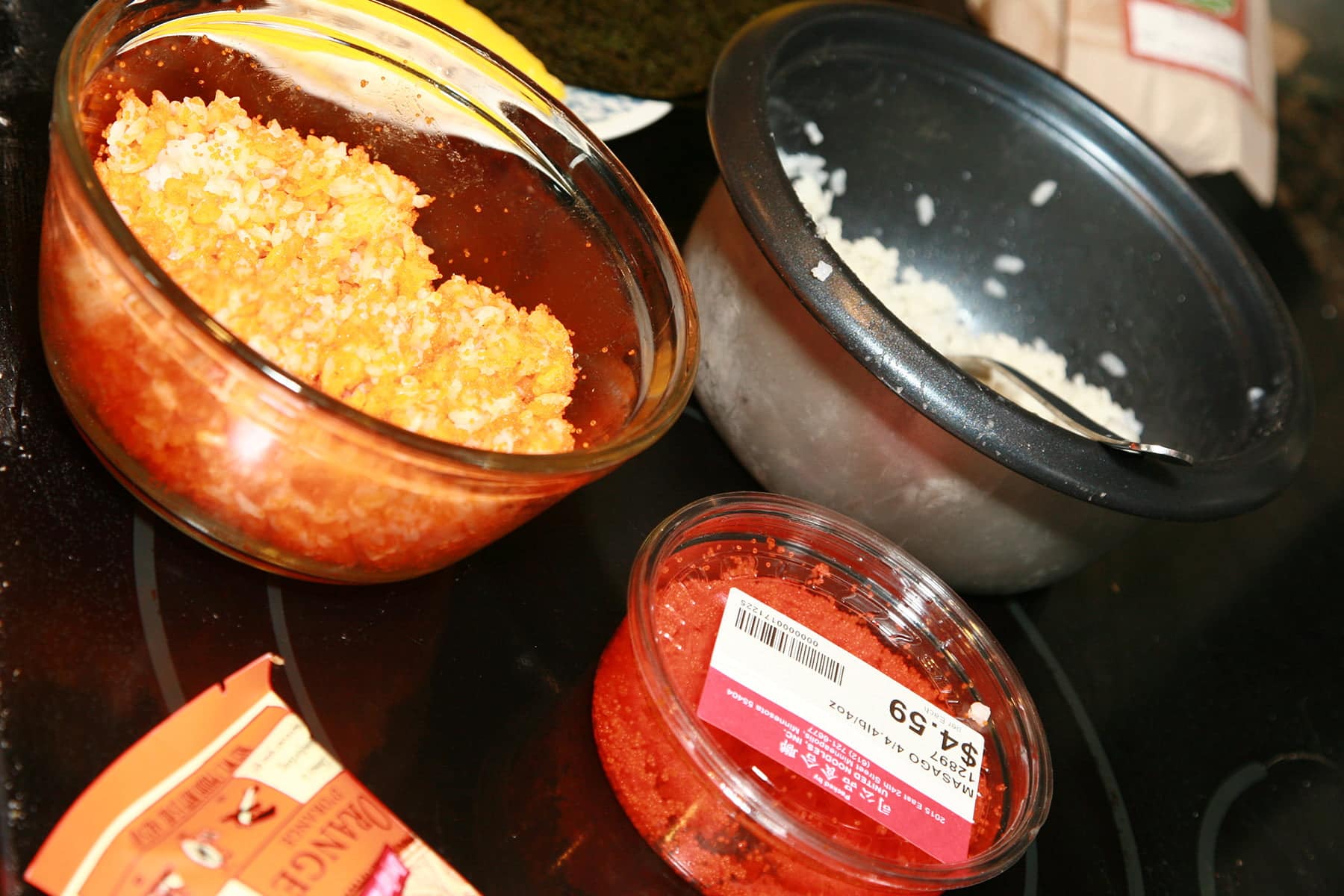 A bowl of orange rice, a rice maker with white rice in it, and a small tub of masago.