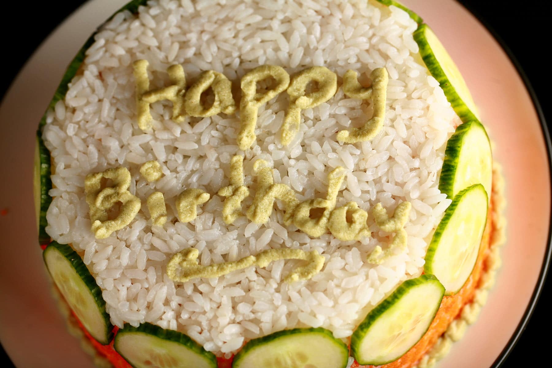 A small "birthday cake" made of white and orange rice and cucumber slices. "Happy Birthday" is written on the top, in wasabi.