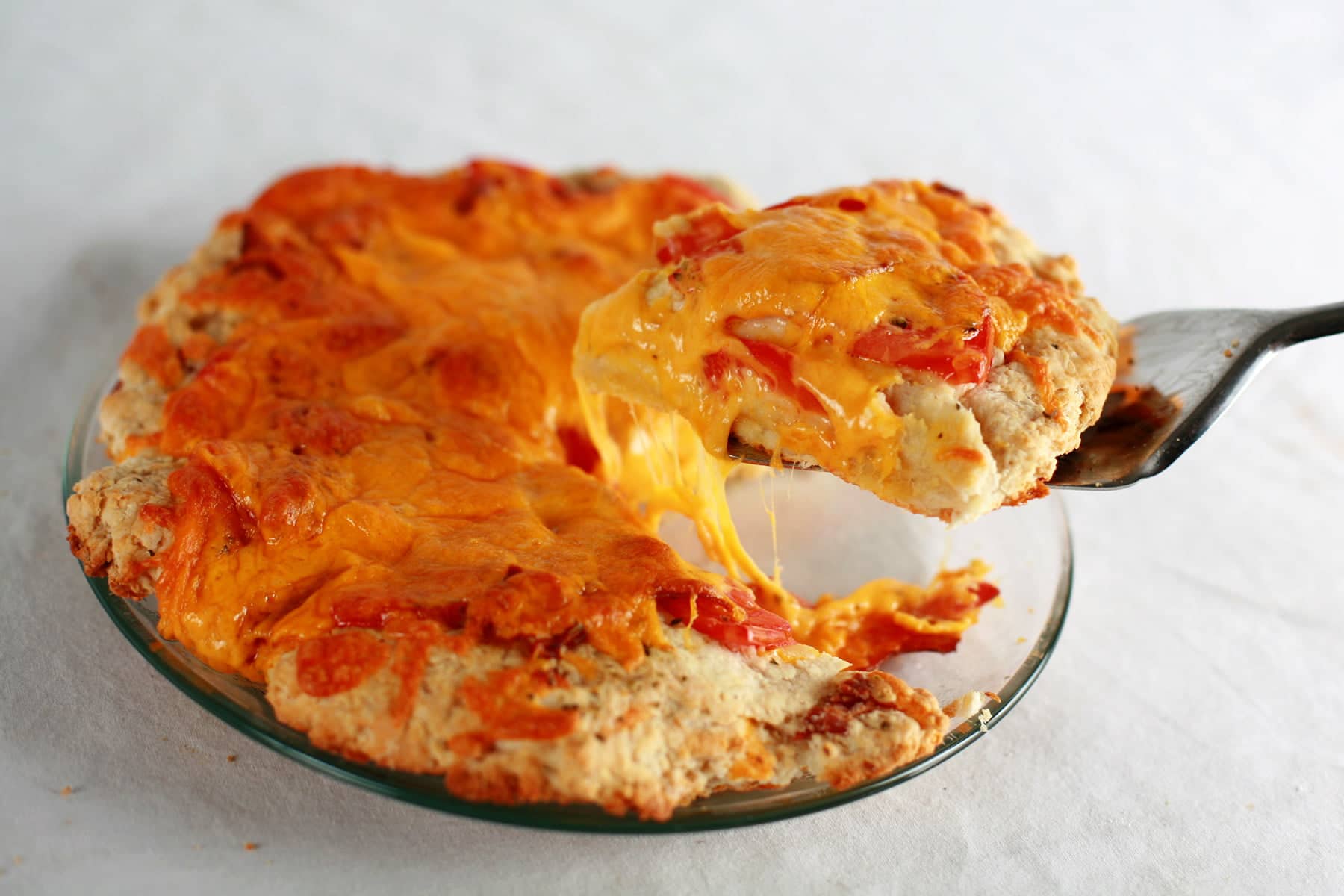 A large round biscuit covered in tomatoes, cheese, and oregano - pizza style - has a wedge cut out of it. A silver coloured spatular is lifting the slice from the rest of the pizza.