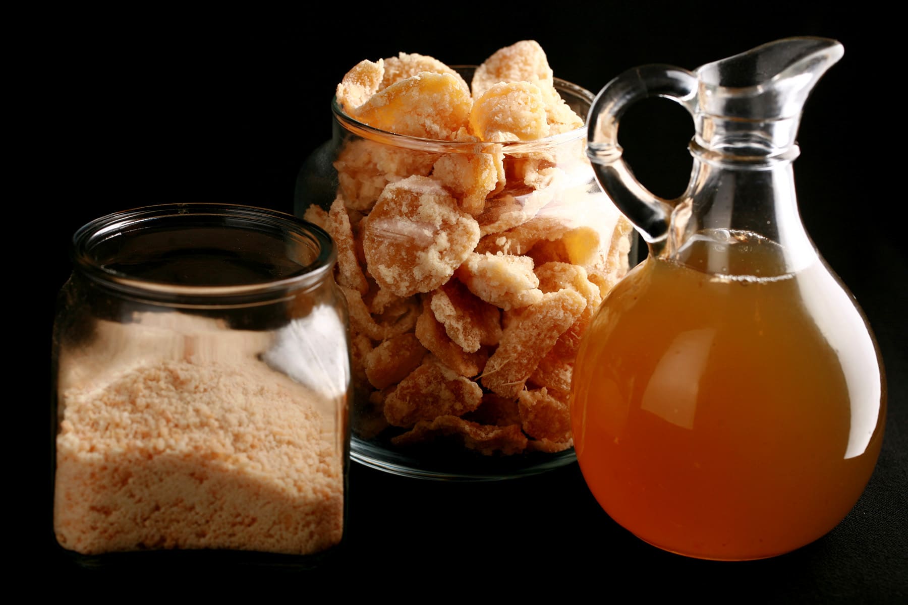 A glass jar of ginger sugar, a taller glass jar filled with candied ginger, and a small bottle of ginger syrup.