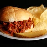 A Sloppy Joes sandwich on a paper plate, with chips and a pickle spear.