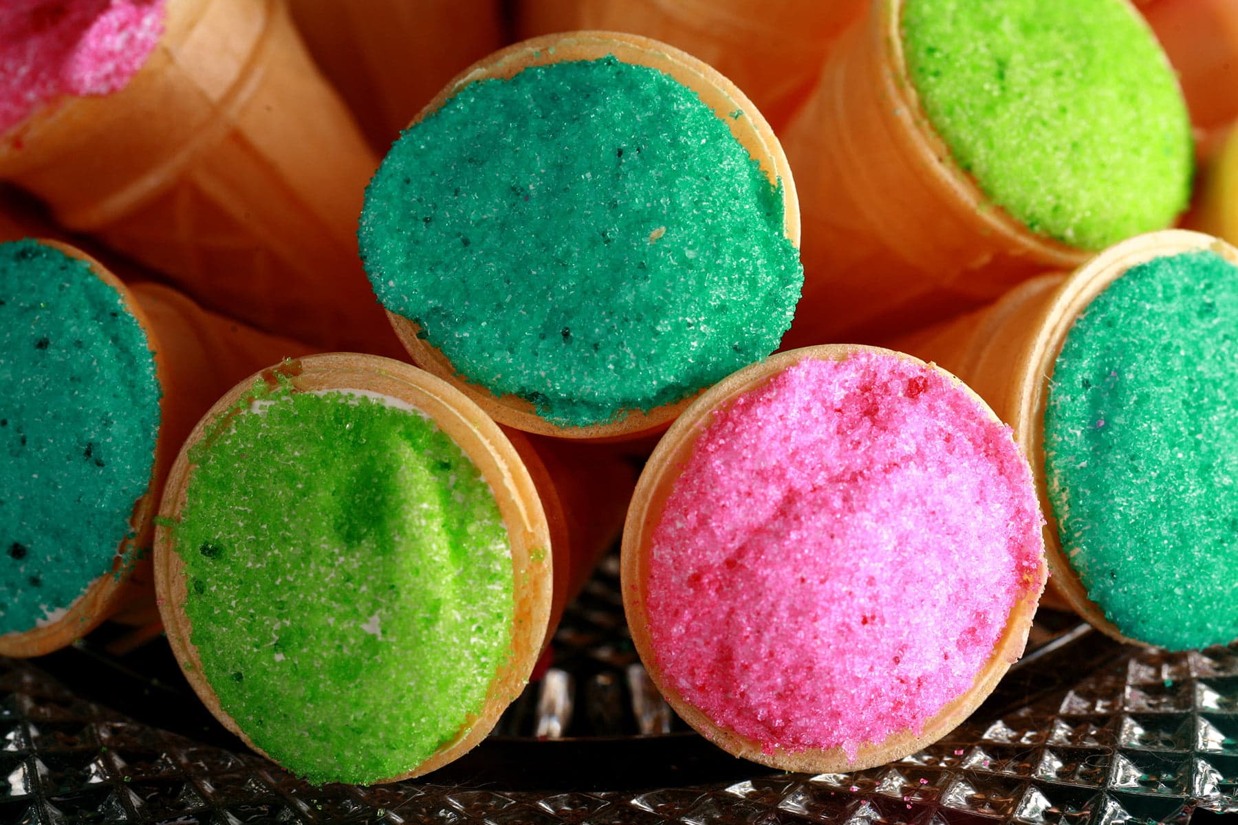 A pile of mini ice cream cones are arranged on a plate. The exposed marshmallow of each is coated in colourful sugar - hot pink, lime green, and teal.