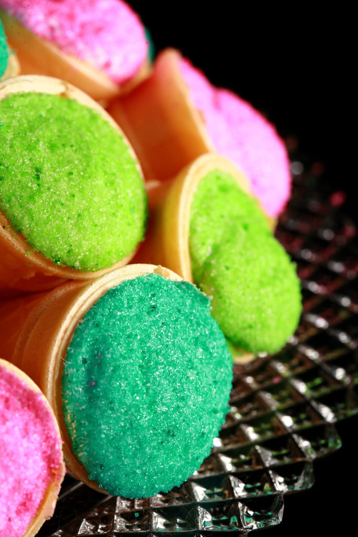 A pile of marshmallow cones - mini ice cream cones filled with marshmallow - are arranged on a plate. The exposed marshmallow of each is coated in colourful sugar - hot pink, lime green, and teal.