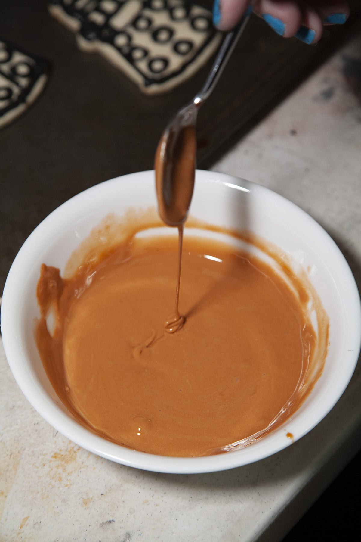 A bowl of brown icing. A spoon drizzles some of it back into the bowl.
