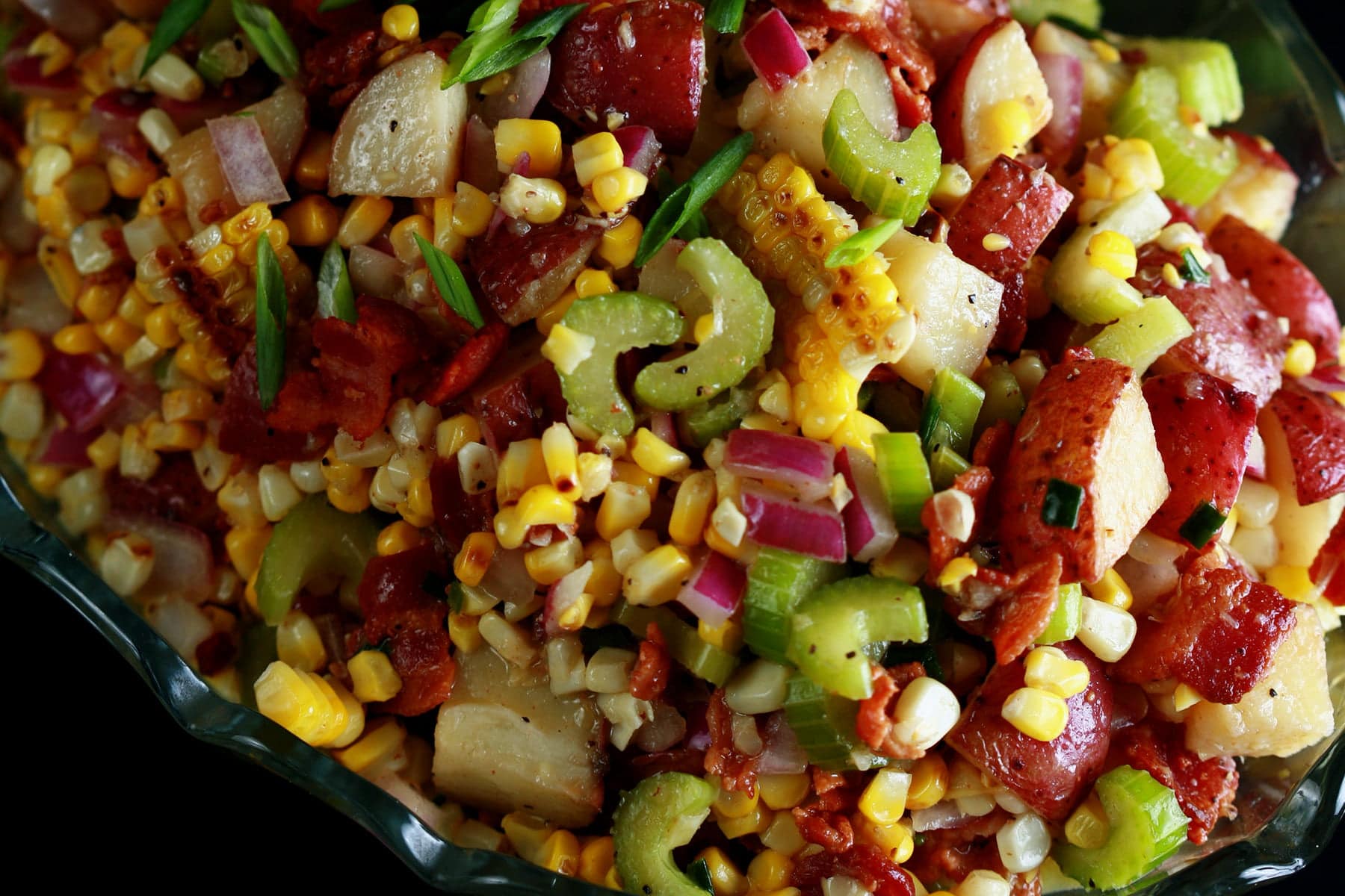 A close up photo of roasted corn and potato salad. Chunks of red potato, bacon pieces, celery slices, green onion, and corn kernels are all visible in the colourful mix.