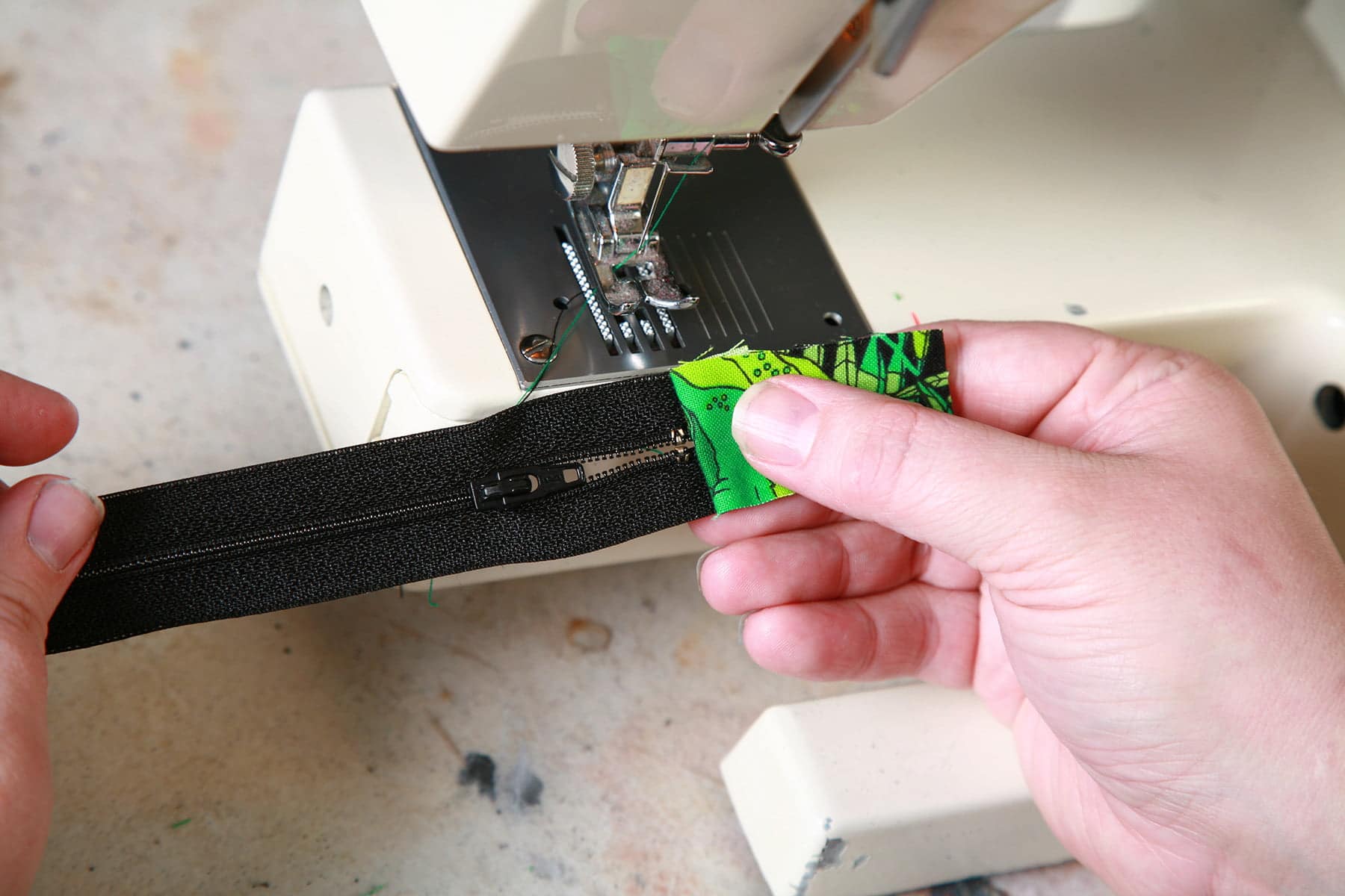 Two hands pull the green fabric back away from the face of a black zipper, holding it in front of a sewing machine.
