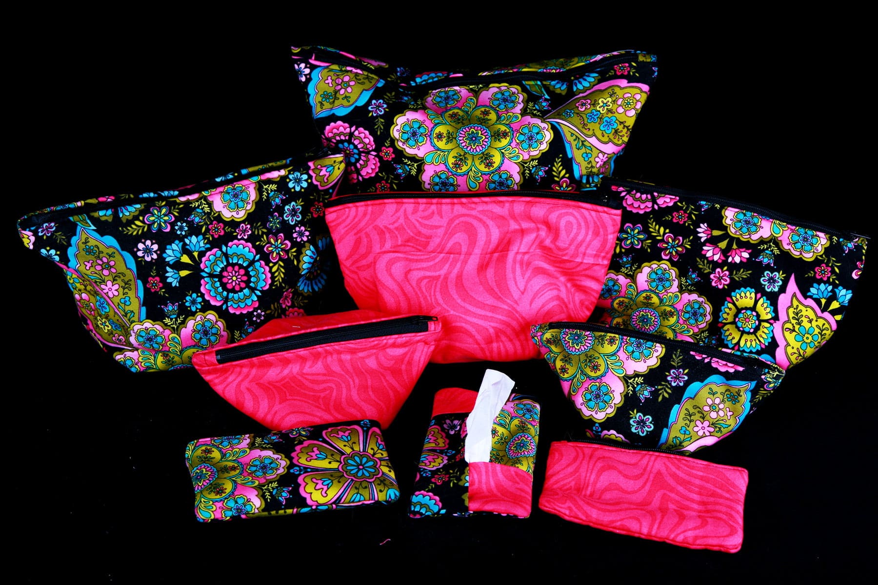 A large travel and makeup bag set - 6 bags of various sizes, two coin purses, and a tissue holder - are shown in front of a black background. The fabrics are a black print with colourful paisley design, and a mottled hot pink.