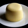 A tall, round serving of sweet corn panna cotta, white plate. The panna cotta is a beautiful butter yellow colour.