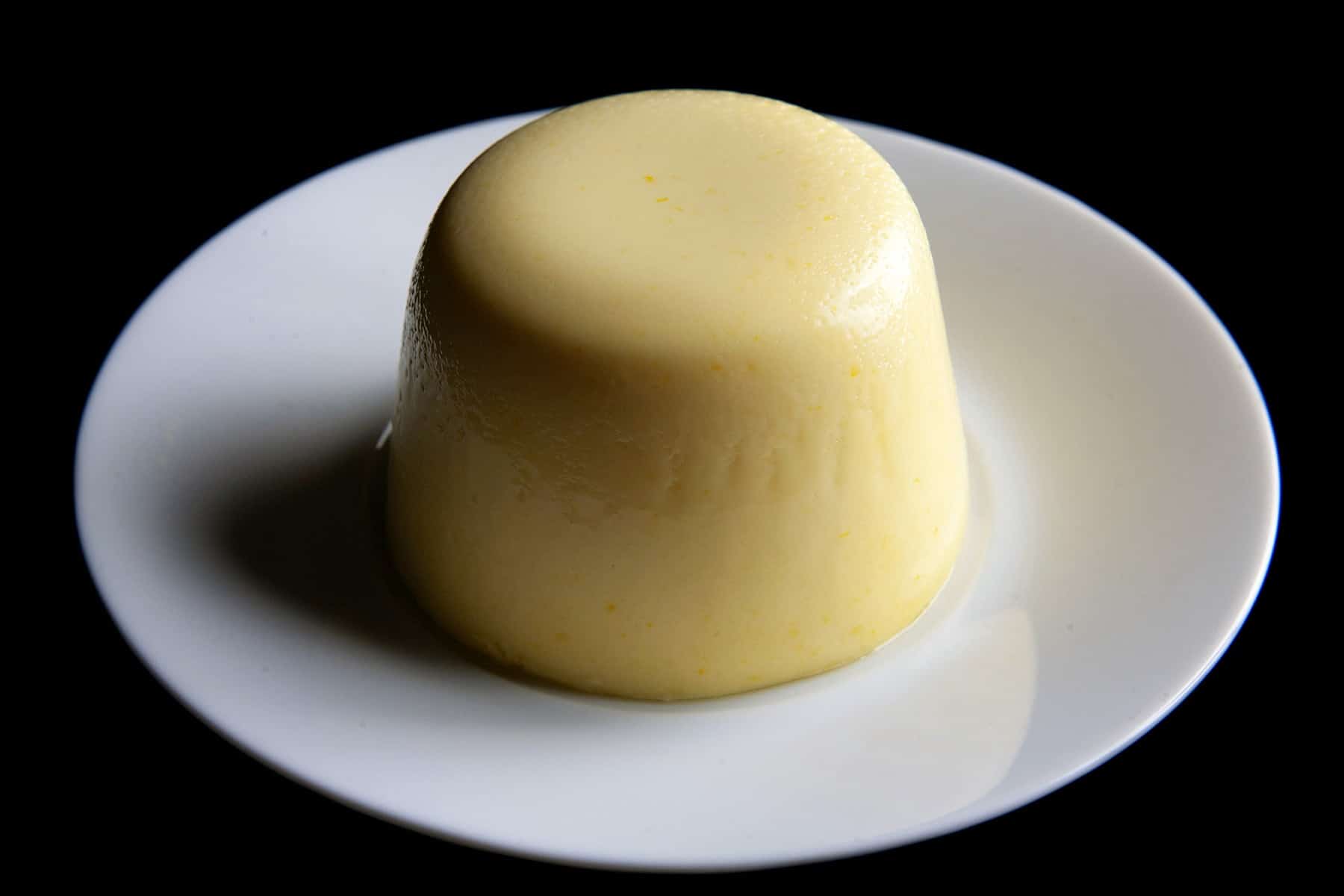 A tall, round serving of sweet corn panna cotta, white plate. The panna cotta is a beautiful butter yellow colour.