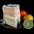 A slice of citrus splendor cake is shown on a plate, with an orange, lemon, and half a lime sitting next to it.