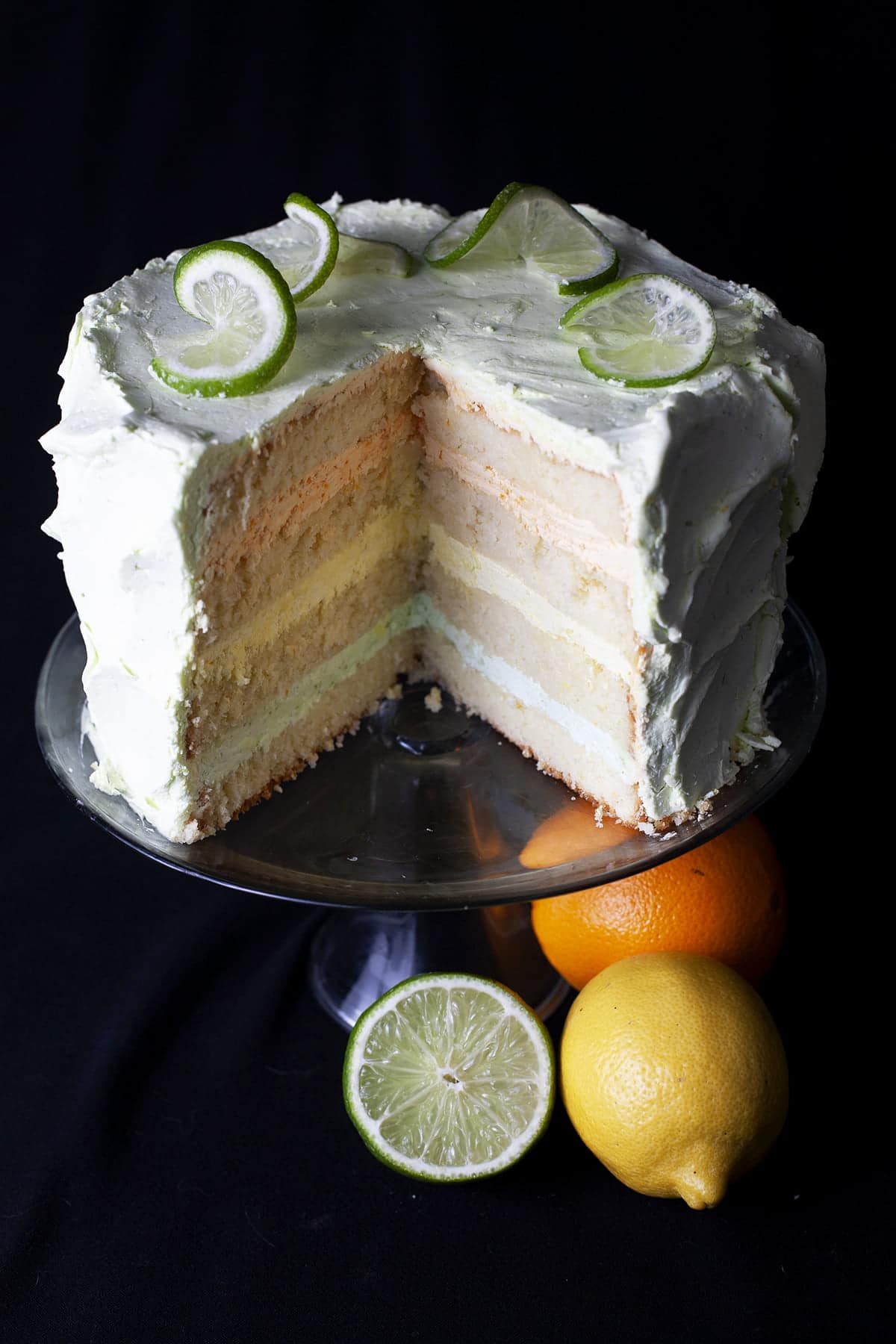 A sliced into citrus splendor cake is shown on a raised plate, with an orange, lemon, and half a lime sitting at the base of the cake stand.