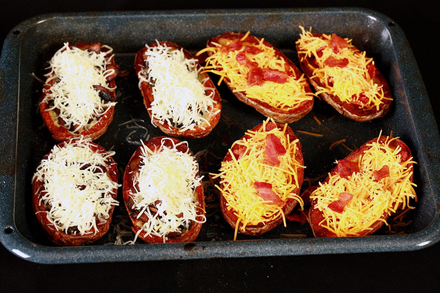 Hollowed out potato skins are arranged on a roasting pan. Half are filled with mushrooms and swiss cheese, the other half have bacon and cheddar as the filling.
