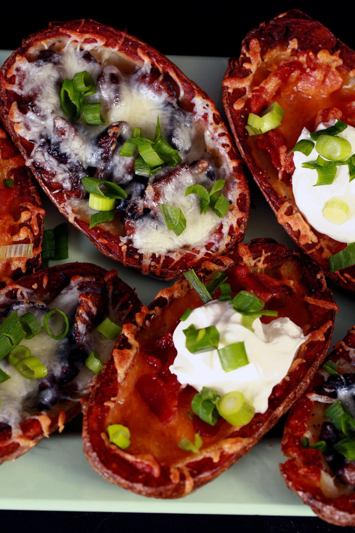 A pale green plate displays 8 bacon roasted potato skins. Half are filled with mushrooms and swiss cheese, the other half have bacon and cheddar as the filling, some topped with sour cream. Most potato skins are sprinkled with sliced green onion.