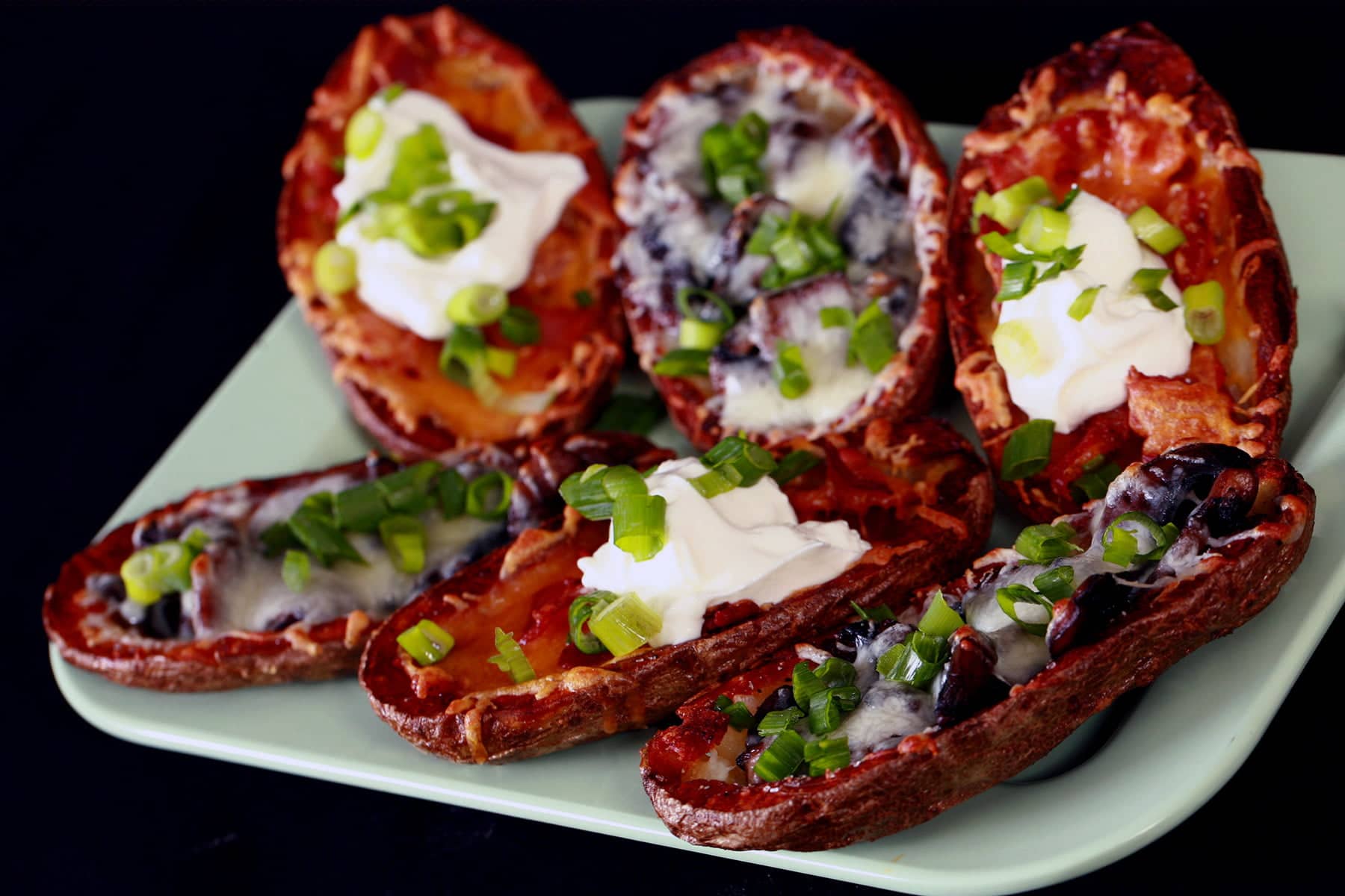 A pale green plate displays 8 baked potato skins. Half are filled with mushrooms and swiss cheese, the other half have bacon and cheddar as the filling, some topped with sour cream. Most potato skins are sprinkled with sliced green onion.