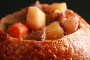 A bread bowl full of hearty beef stew. Chunks of beef, carrots, and potatoes are prominent.