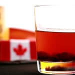 A tumbler glass of maple butter tart liqueur, next to a bottle of maple syrip and a small Canadian flag.