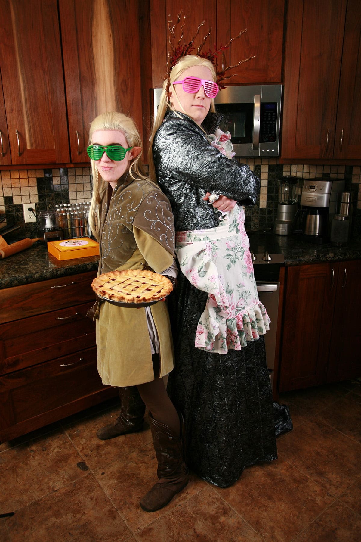 Cosplayers dressed as Thranduil and Legolas are back to back in the kitchen. Legolas is holding a pie, and both are wearing 80s style slat sunglasses.