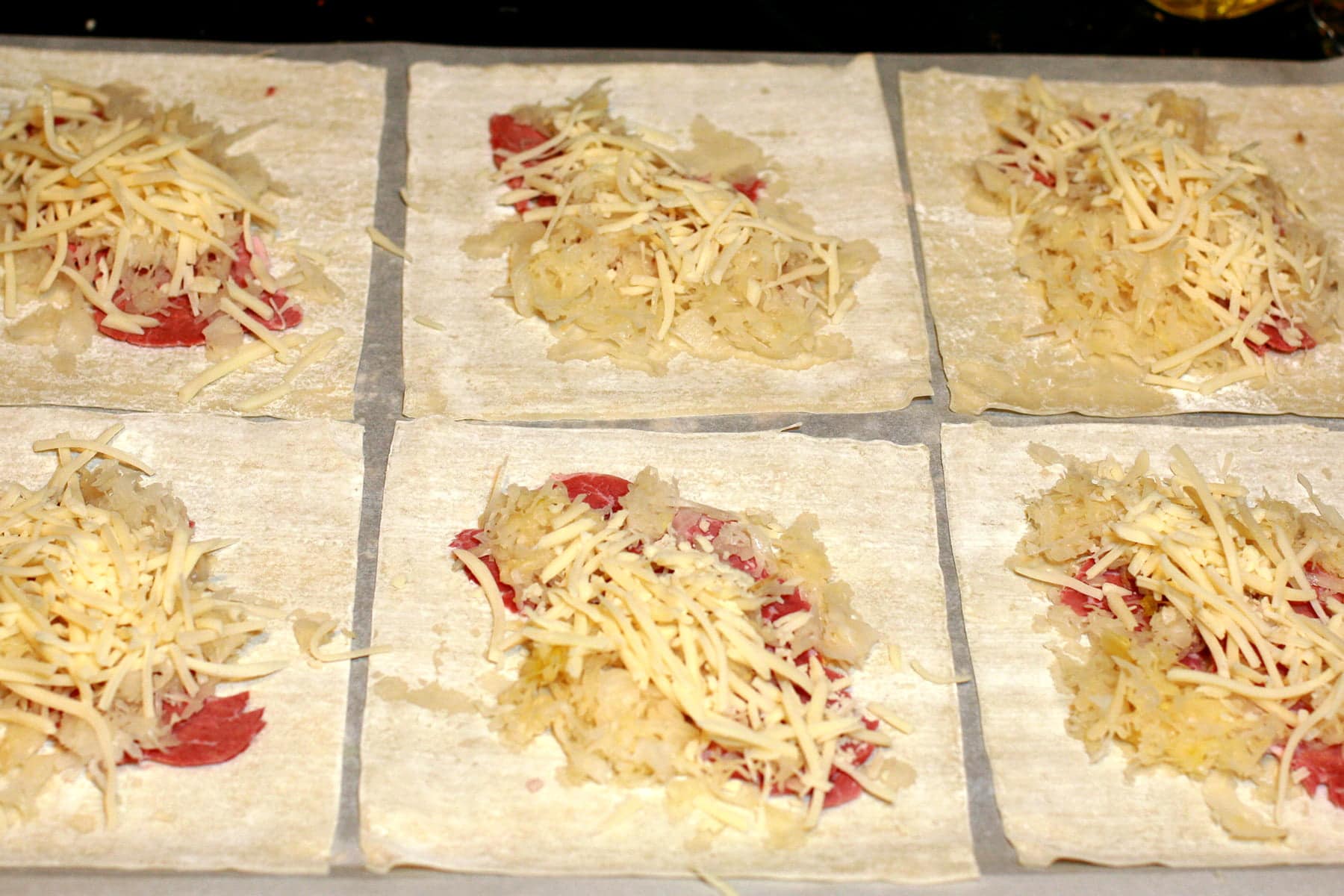 Wonton wrappers are arranged on a work surface. Each has meat, sauerkraut, and swiss cheese on top.