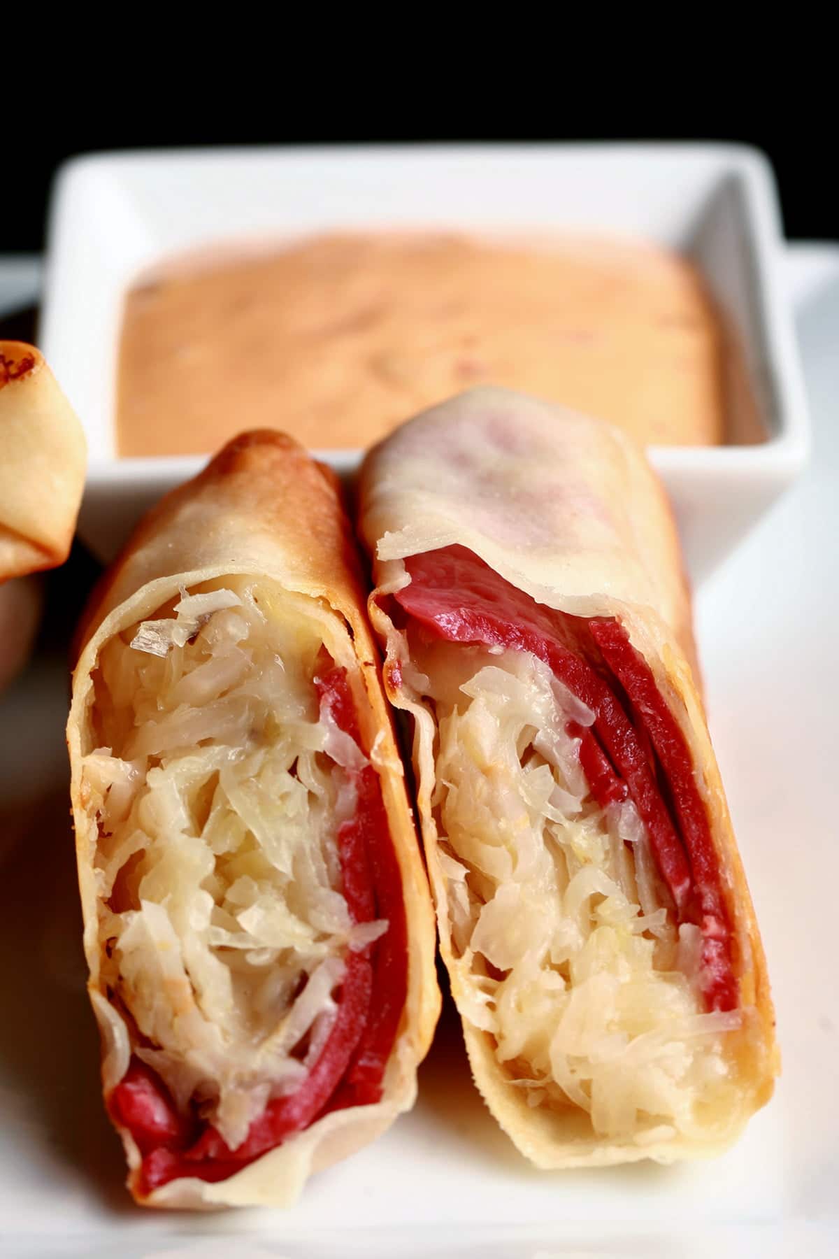 A white plate is piled with baked Reuben egg rolls, with one cut in half. There is a small bowl of thousand island dressing on the plate, as well.