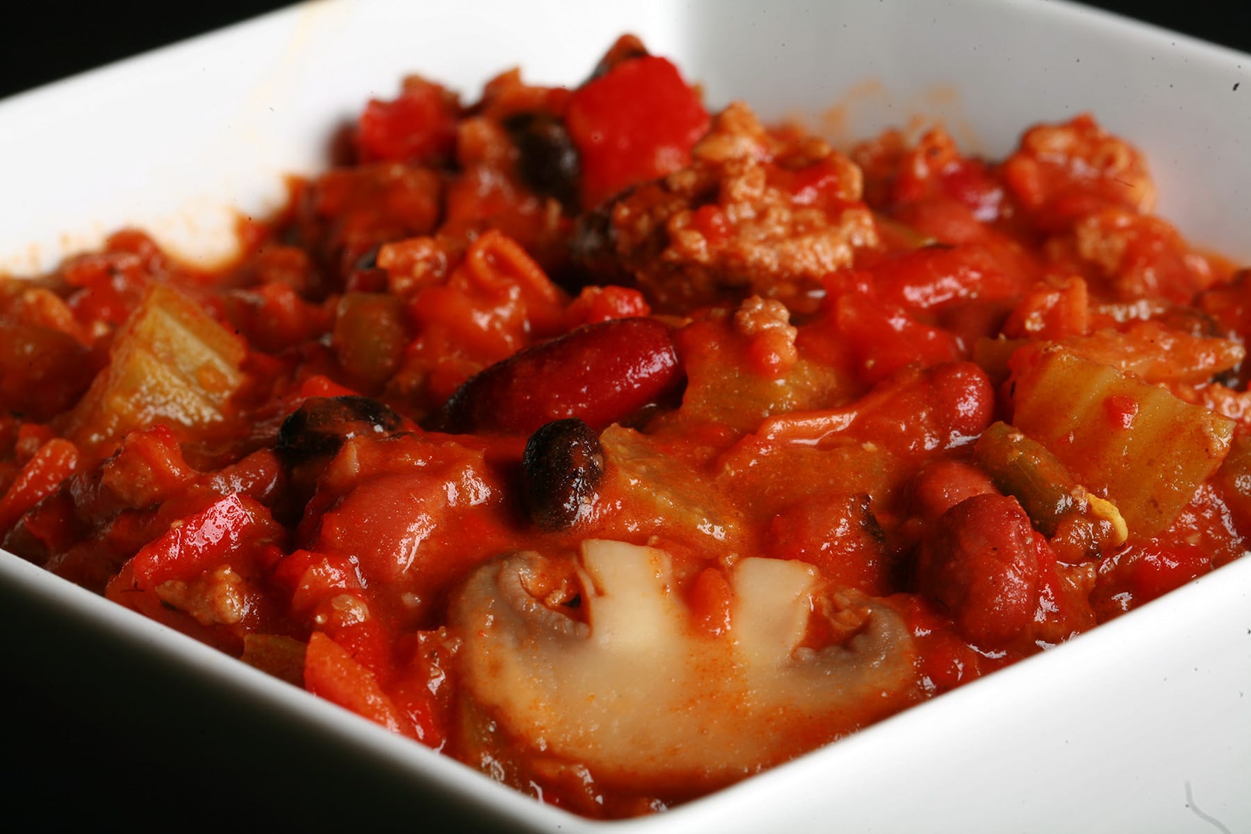 Close up photo of a bowl of roasted convention chili. Kidney beans, ground beef, mushrooms, celery, and red peppers are visible.
