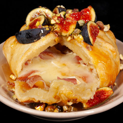 A baked brie en croute, wrapped in puff pastry, with prosciutto, figs, honey pear sauce, and pistachios.