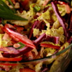 A glass bowl is filled with a colourful coleslaw. Red pepper, purple and green cabbage, carrot, and green onions are all visible.